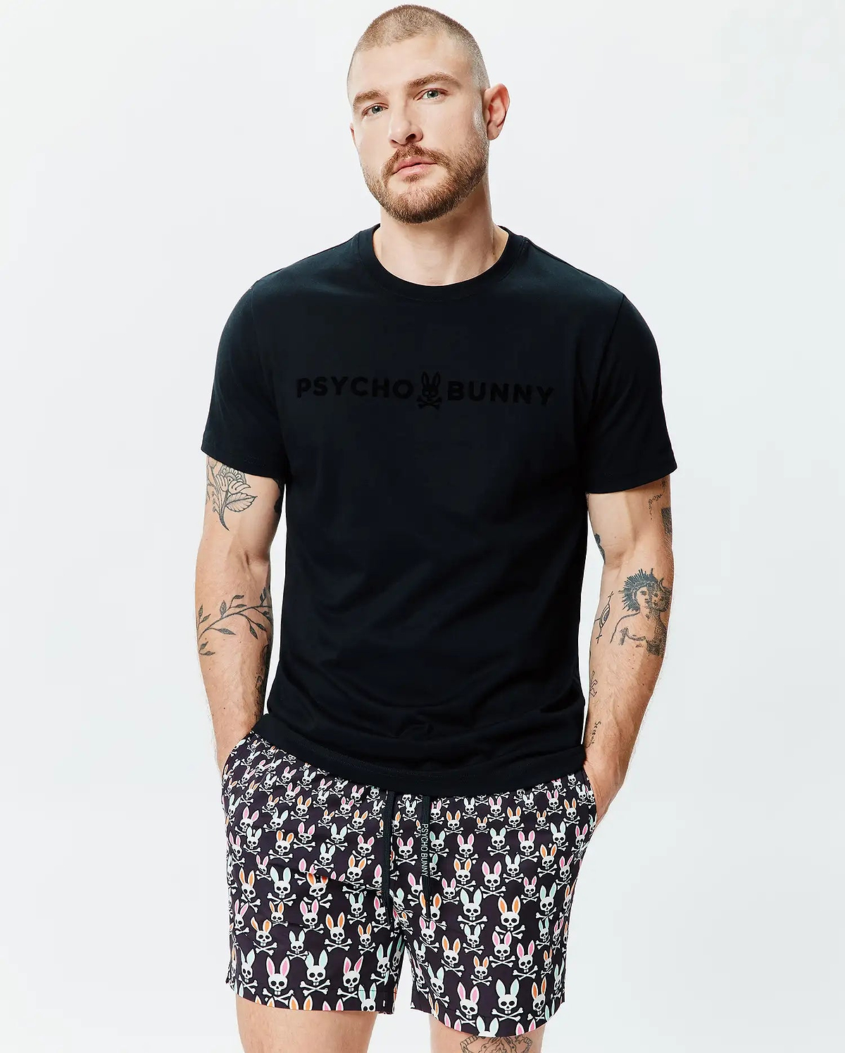 Mens Royce Graphic Tee, a black jersey t-shirt with a tone-on-tone PSYCHO BUNNY logo on the chest, paired with black shorts featuring a multi-colored bunny skull pattern. (2)