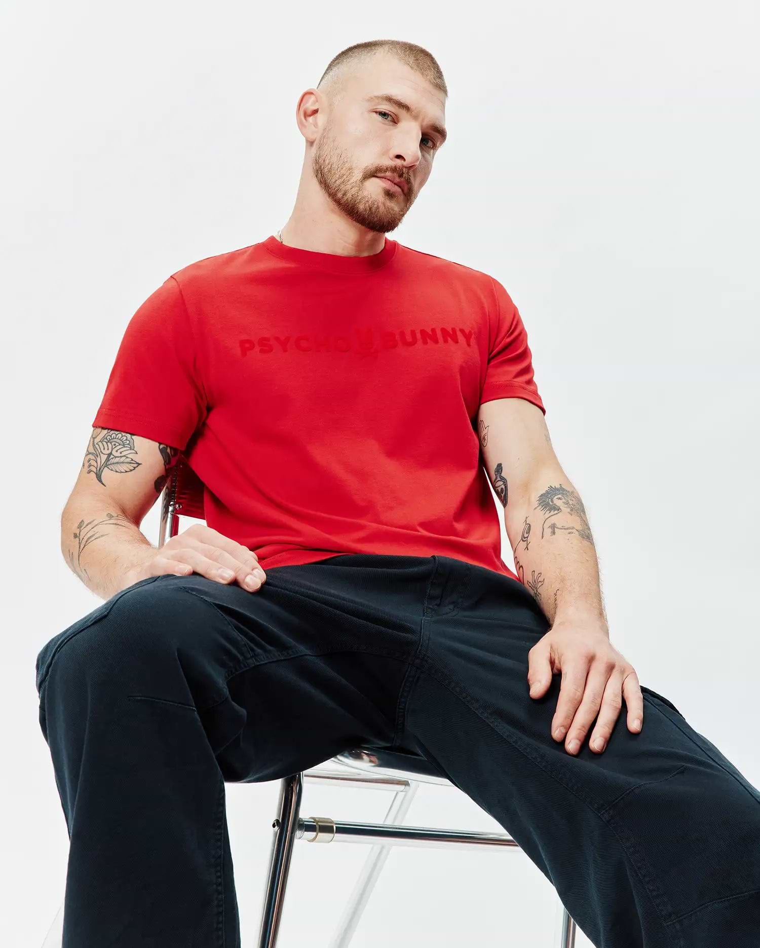 Mens Royce Graphic Tee: Red classic fit jersey t-shirt with a large, embossed red PSYCHO BUNNY chest logo, complemented by relaxed dark blue utility pants.