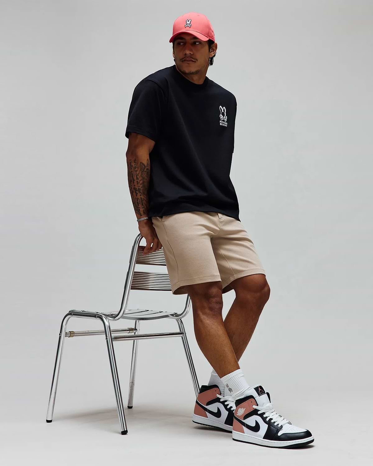 Black Mens Toby Oversized Tee in jersey knit with white Psycho Bunny skull logo and text on left chest, paired with khaki shorts and a coral cap featuring a white embroidered Psycho Bunny skull logo. (3)