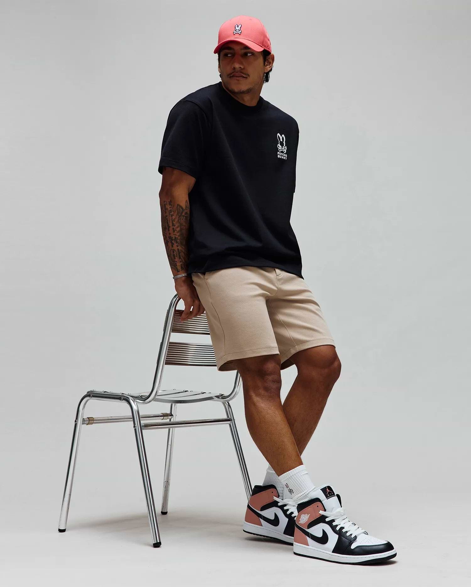 Black Mens Toby Oversized Tee in jersey knit with white Psycho Bunny skull logo and text on left chest, paired with khaki shorts and a coral cap featuring a white embroidered Psycho Bunny skull logo. (3)