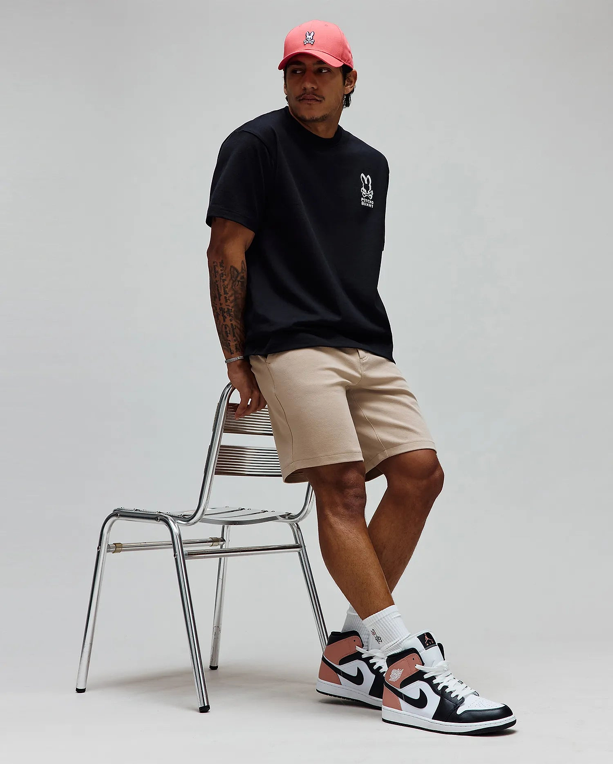 Black Mens Toby Oversized Tee in jersey knit with white Psycho Bunny skull logo and text on left chest, paired with khaki shorts and a coral cap featuring a white embroidered Psycho Bunny skull logo. (3)