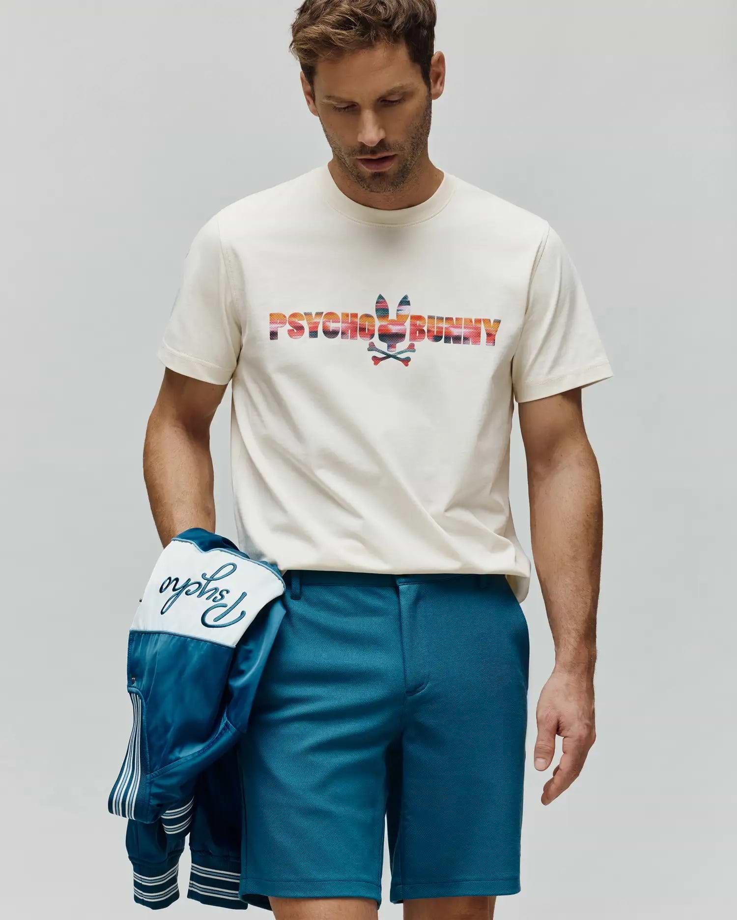 Mens Raphael Wordmark Tee, an off-white jersey tee with a large, colorful striped Psycho Bunny wordmark and bunny skull chest logo. Teal shorts and a satin jacket with embroidered Psycho script. (1)