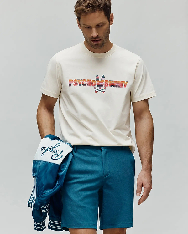 Mens Raphael Wordmark Tee, an off-white jersey tee with a large, colorful striped Psycho Bunny wordmark and bunny skull chest logo. Teal shorts and a satin jacket with embroidered Psycho script.