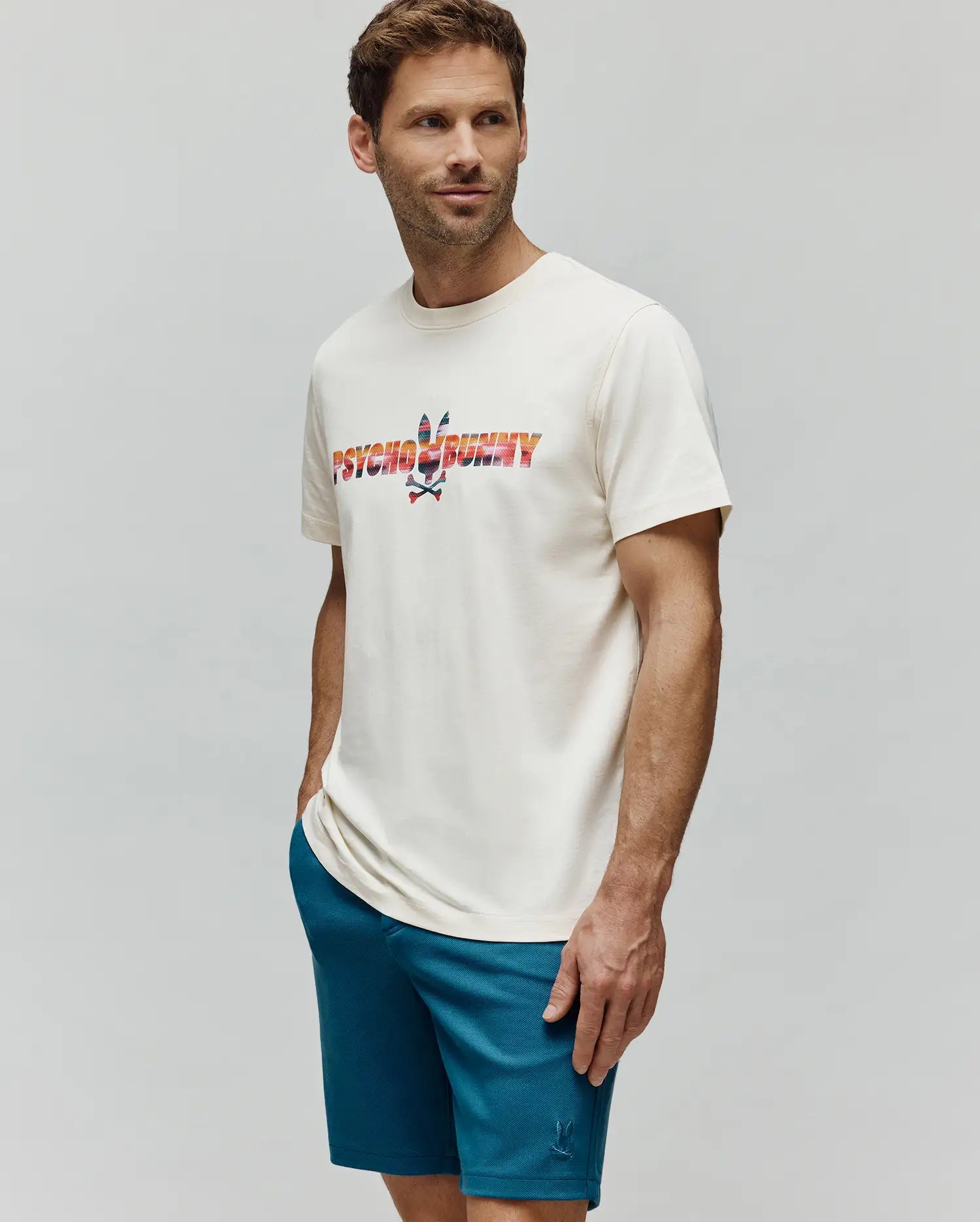 Mens Raphael Wordmark Tee, a cream classic fit jersey t-shirt with a large, vibrant multi-color gradient Psycho Bunny wordmark and skull logo chest graphic. Paired with teal textured knit shorts featuring a small embroidered bunny logo. (3)