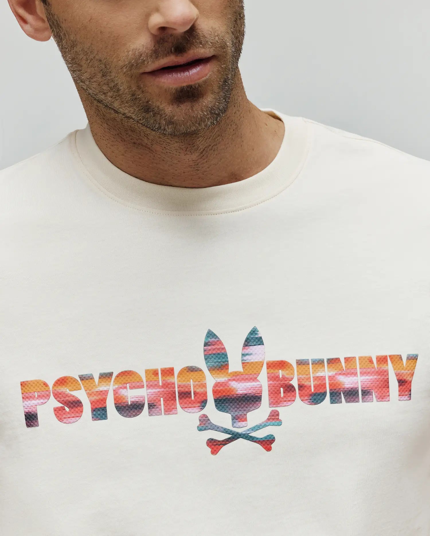 Mens Raphael Wordmark Tee, a cream jersey knit t-shirt featuring a large center chest graphic of the Psycho Bunny wordmark and iconic bunny skull logo in a vibrant multi-color striped pattern. (2)