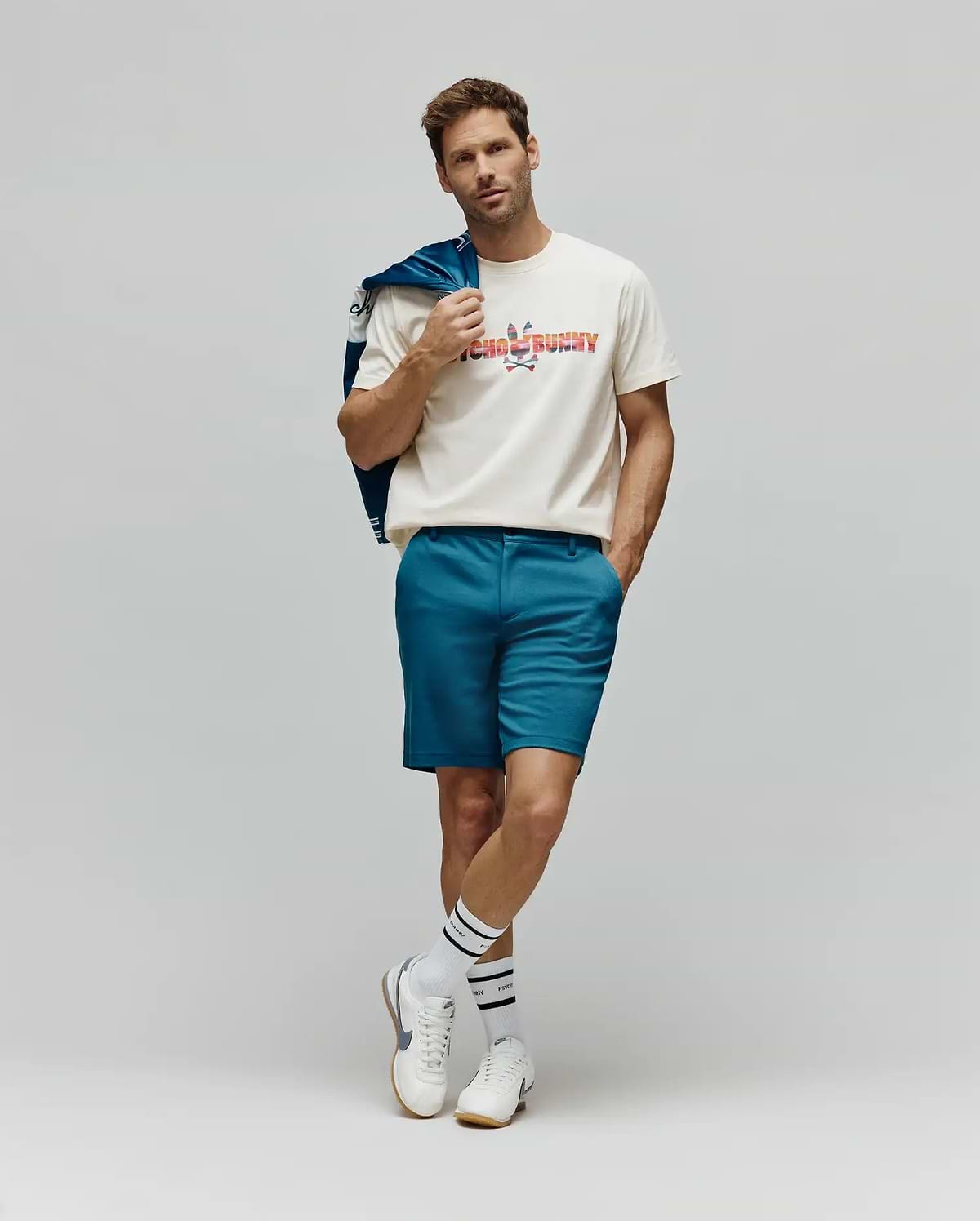 Mens Raphael Wordmark Tee, a classic fit cream jersey t-shirt featuring a large, multicolored patterned Psycho Bunny wordmark and skull logo on the chest, complemented by slim-fit teal shorts. (4)