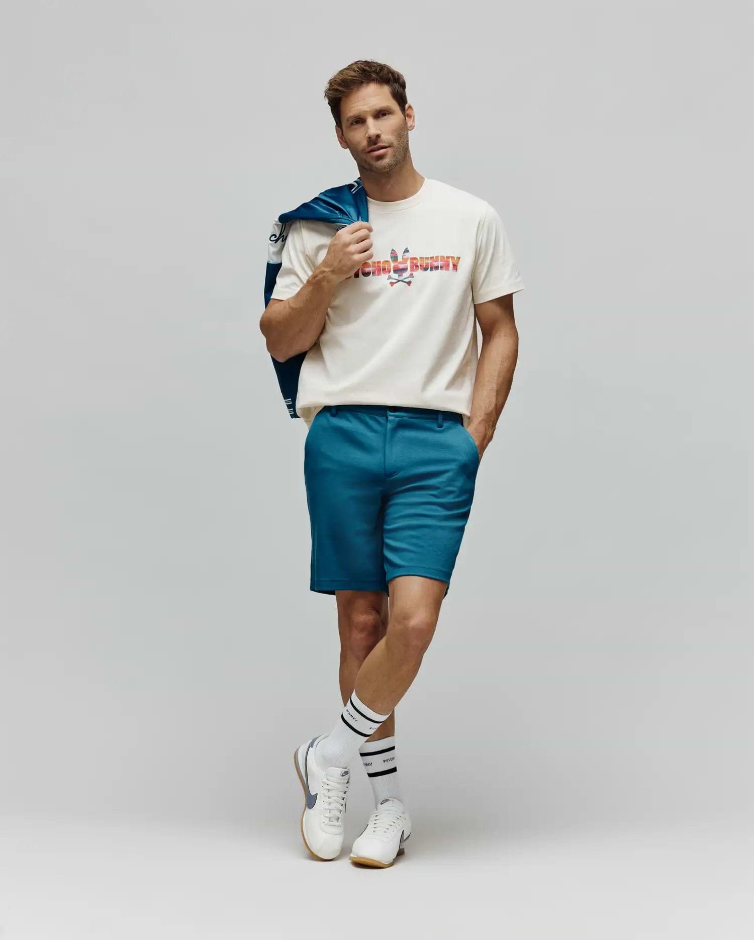 Mens Raphael Wordmark Tee, a classic fit cream jersey t-shirt featuring a large, multicolored patterned Psycho Bunny wordmark and skull logo on the chest, complemented by slim-fit teal shorts. (4)
