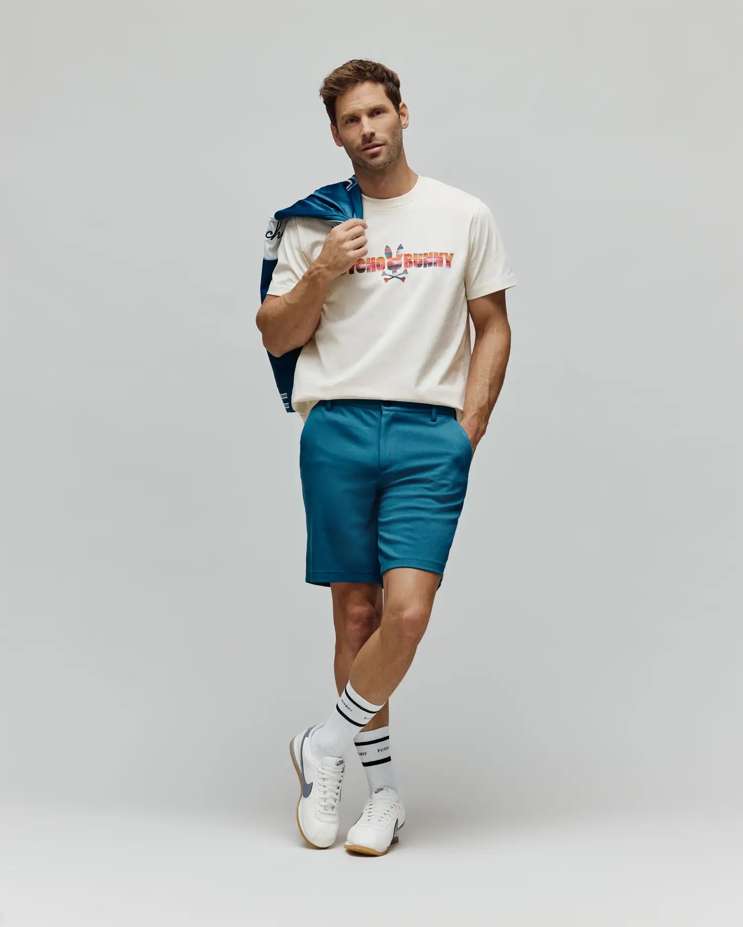 Mens Raphael Wordmark Tee, a classic fit cream jersey t-shirt featuring a large, multicolored patterned Psycho Bunny wordmark and skull logo on the chest, complemented by slim-fit teal shorts. (4)