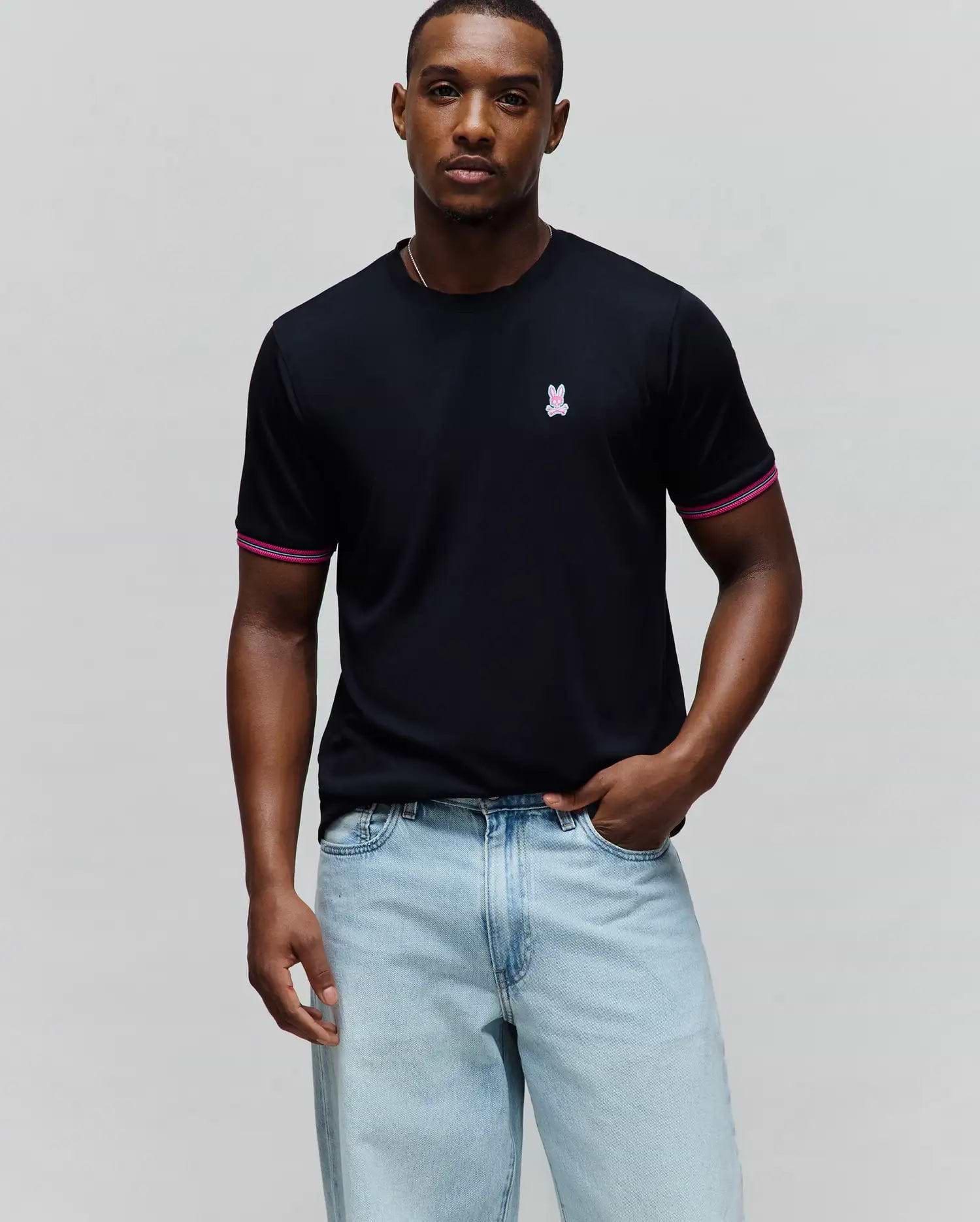 Mens Clark Performance Tee, black jersey knit with a classic fit, featuring a small embroidered pink and white Psycho Bunny skull logo on the left chest and striped pink and white cuffs.