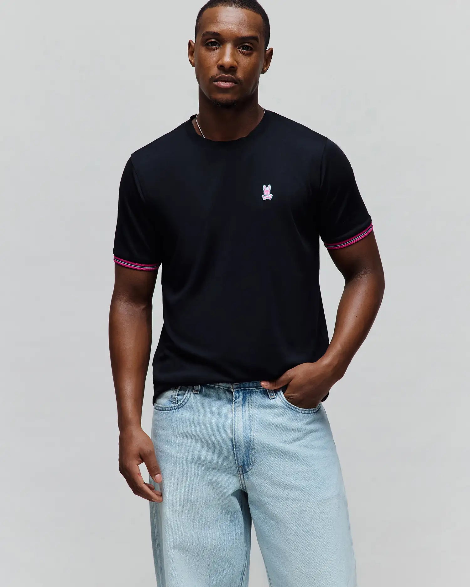 Mens Clark Performance Tee, black jersey knit with a classic fit, featuring a small embroidered pink and white Psycho Bunny skull logo on the left chest and striped pink and white cuffs.