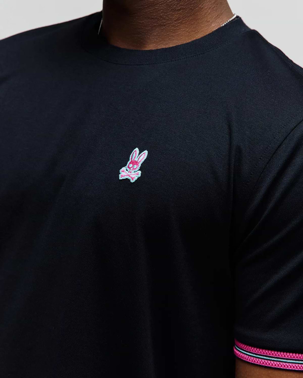 Mens Clark Performance Tee, black classic fit smooth jersey knit with a small embroidered pink and light blue Psycho Bunny skull logo on the left chest and striped sleeve trim.
