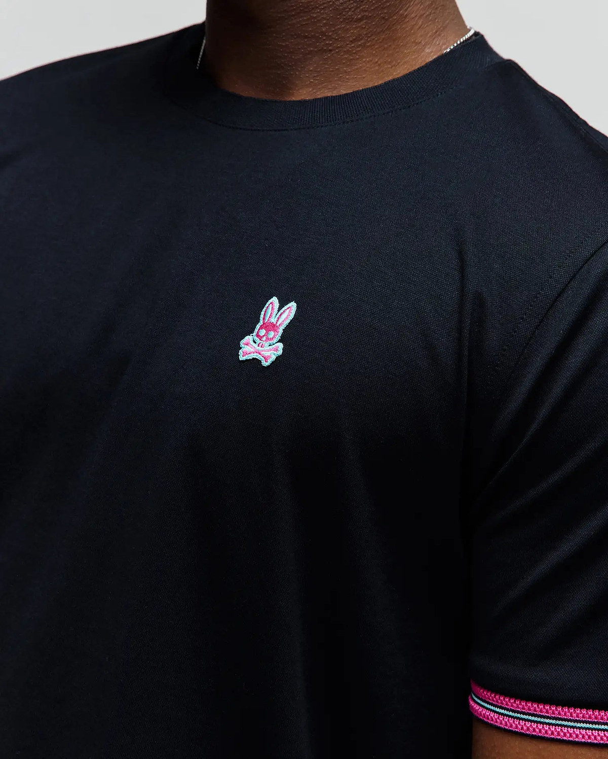 Mens Clark Performance Tee, black classic fit smooth jersey knit with a small embroidered pink and light blue Psycho Bunny skull logo on the left chest and striped sleeve trim.