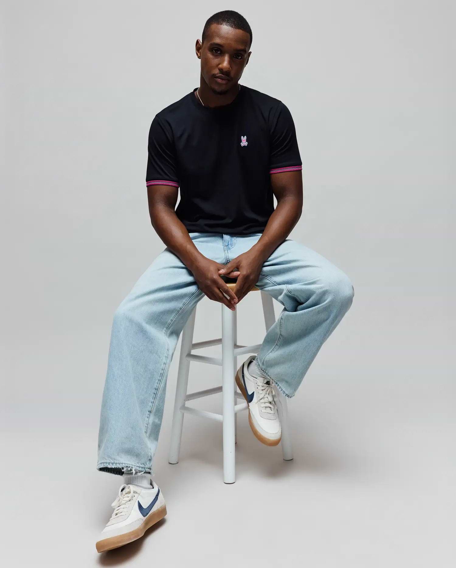Mens Clark Performance Tee, a black classic fit jersey t-shirt with a small embroidered pink Psycho Bunny skull logo on the left chest and pink striped sleeve cuffs.