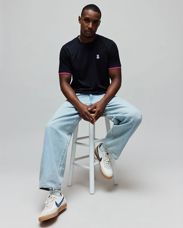 Mens Clark Performance Tee, a black classic fit jersey t-shirt with a small embroidered pink Psycho Bunny skull logo on the left chest and pink striped sleeve cuffs.