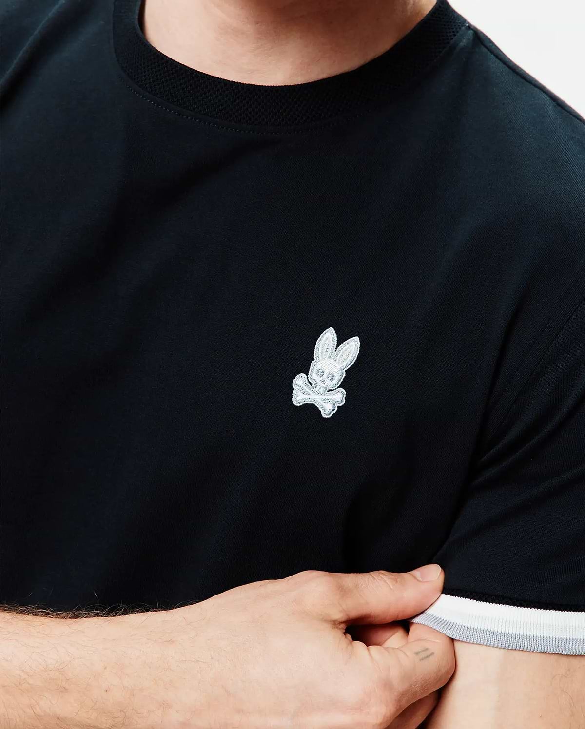 Mens Holden Tee, black jersey knit with a ribbed crew neck, featuring a small embroidered white Psycho Bunny skull logo on the left chest and ribbed cuffs with white and grey stripes.
