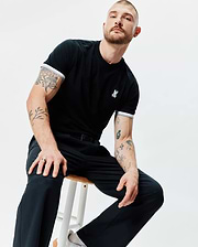 Black Mens Holden Tee in classic fit jersey knit, featuring a small embroidered white Psycho Bunny skull logo on the left chest and white and grey striped ribbed sleeve cuffs.