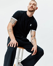 Black Mens Holden Tee in classic fit jersey knit, featuring a small embroidered white Psycho Bunny skull logo on the left chest and white and grey striped ribbed sleeve cuffs.