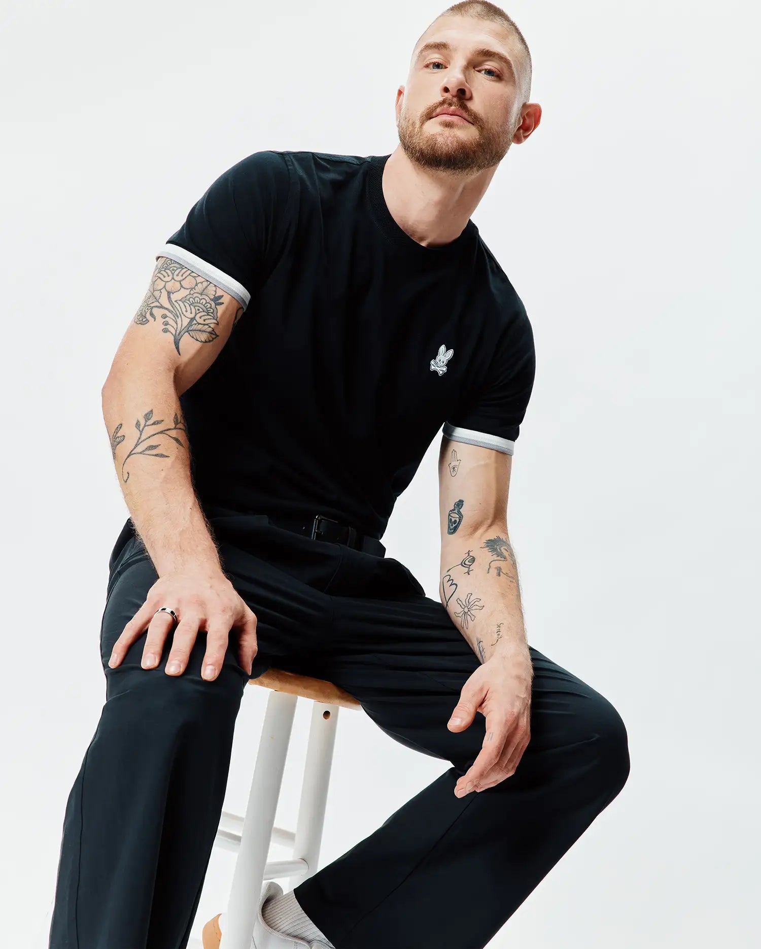 Black Mens Holden Tee in classic fit jersey knit, featuring a small embroidered white Psycho Bunny skull logo on the left chest and white and grey striped ribbed sleeve cuffs.