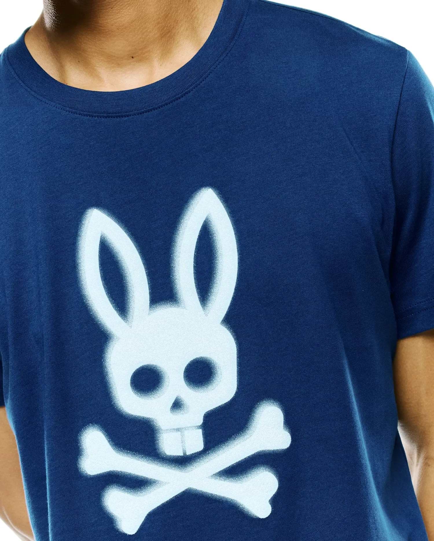 Mens Emmanuel Graphic Tee, a classic fit navy blue jersey t-shirt featuring a large, light blue spray-painted Psycho Bunny skull and crossbones graphic on the center chest.