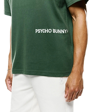 Mens Haiden Relaxed Heavy Weight Tee in dark green heavyweight jersey, featuring a large white embroidered Psycho Bunny text logo on the chest, paired with white textured shorts displaying a small bunny skull logo.