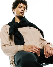 Mens Norman Long Sleeve Relaxed Fit Tee in beige jersey, featuring a small embroidered black Psycho Bunny skull logo on the left chest, paired with black pants and a black sweater tied over the shoulders.