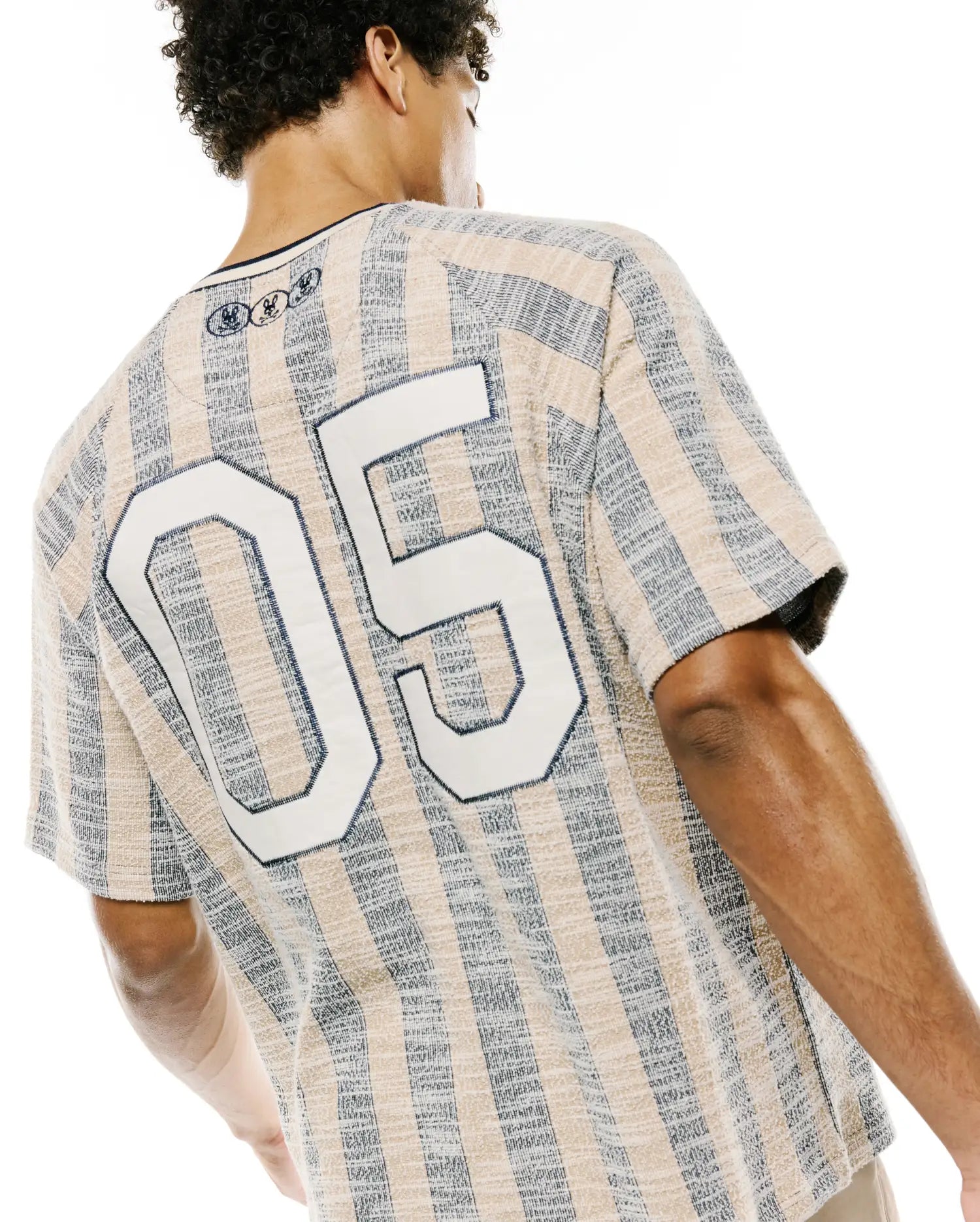 Mens Nathaniel Relaxed Raglan Tee in textured beige and navy vertical stripes, featuring a large white 05 graphic on the back and three small embroidered Psycho Bunny skull logos at the upper back neckline.