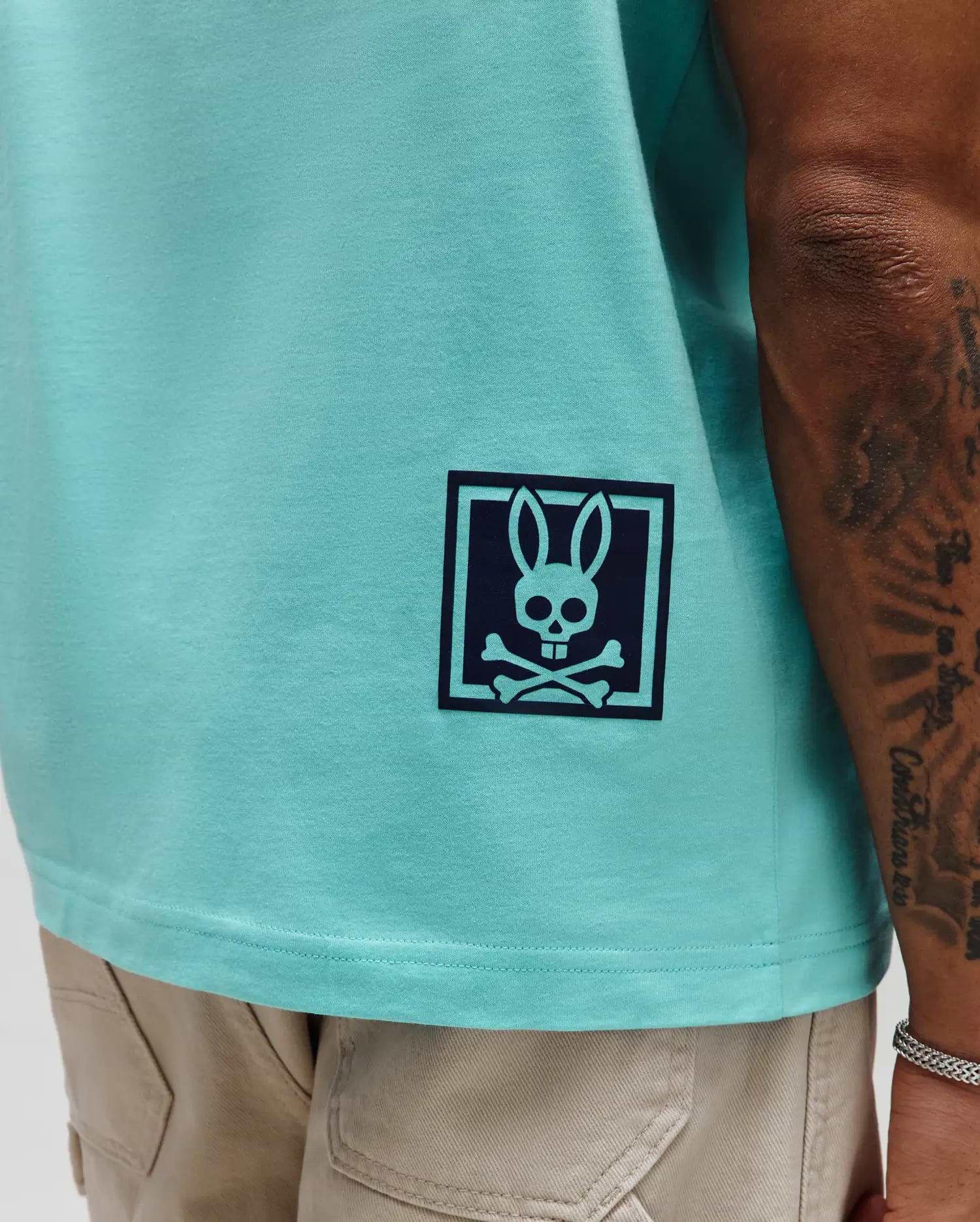Mens Keon Graphic Tee, aqua smooth jersey knit, classic fit, with a dark blue Psycho Bunny skull and crossbones graphic in a square frame on the lower left side.