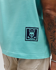 Mens Keon Graphic Tee, aqua smooth jersey knit, classic fit, with a dark blue Psycho Bunny skull and crossbones graphic in a square frame on the lower left side.