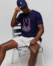 Mens Keon Embroidered Graphic Tee in navy blue, classic-fit, with a large pink Psycho Bunny skull graphic, paired with light beige drawstring shorts and a navy cap featuring an embroidered bunny. (4)