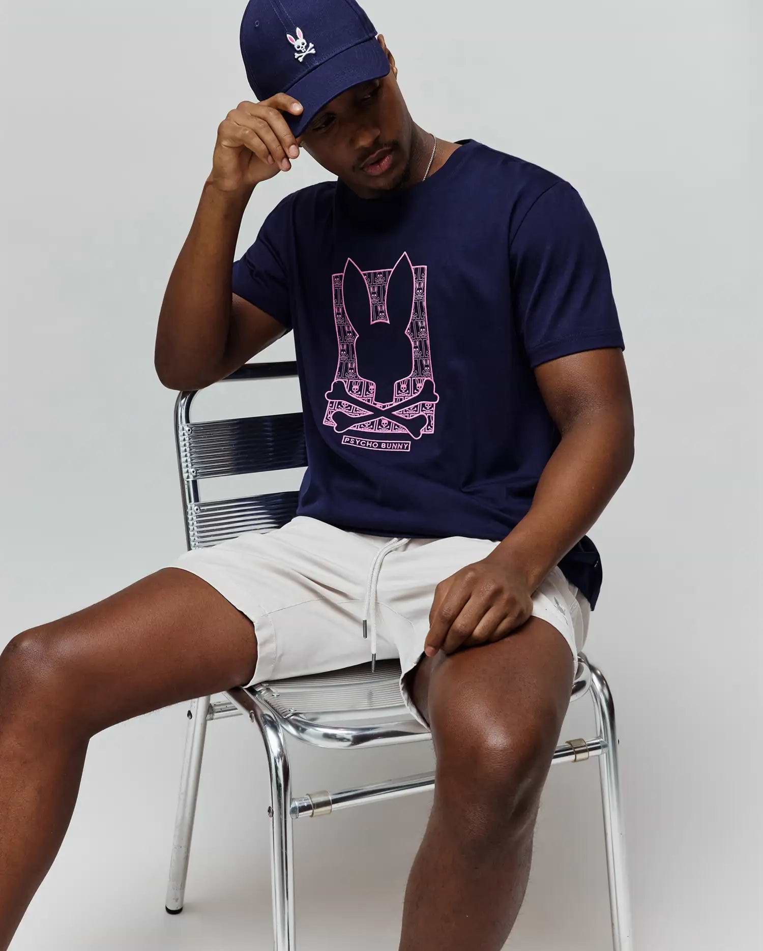 Mens Keon Embroidered Graphic Tee in navy blue, classic-fit, with a large pink Psycho Bunny skull graphic, paired with light beige drawstring shorts and a navy cap featuring an embroidered bunny. (4)