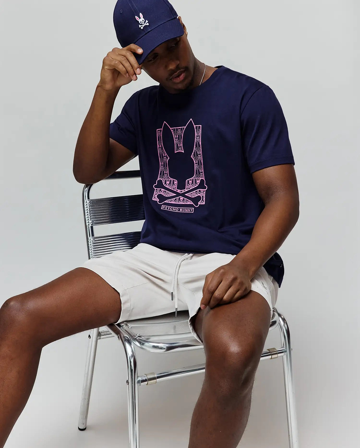 Mens Keon Embroidered Graphic Tee in navy blue, classic-fit, with a large pink Psycho Bunny skull graphic, paired with light beige drawstring shorts and a navy cap featuring an embroidered bunny. (4)