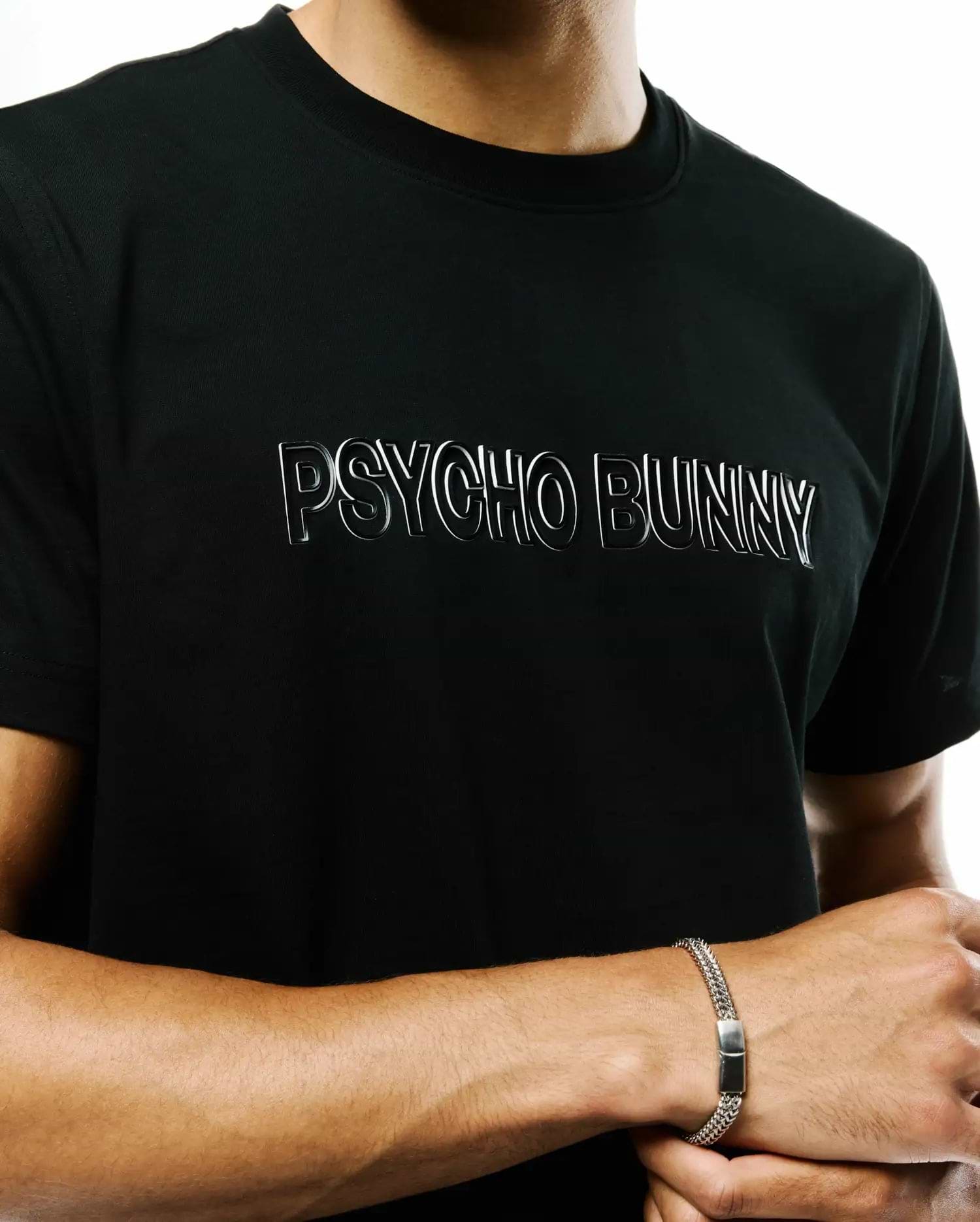 Mens Joji Wordmark Graphic Tee, a black classic fit jersey knit t-shirt featuring a prominent silver outlined Psycho Bunny wordmark graphic on the chest and a small bunny skull logo on the sleeve.