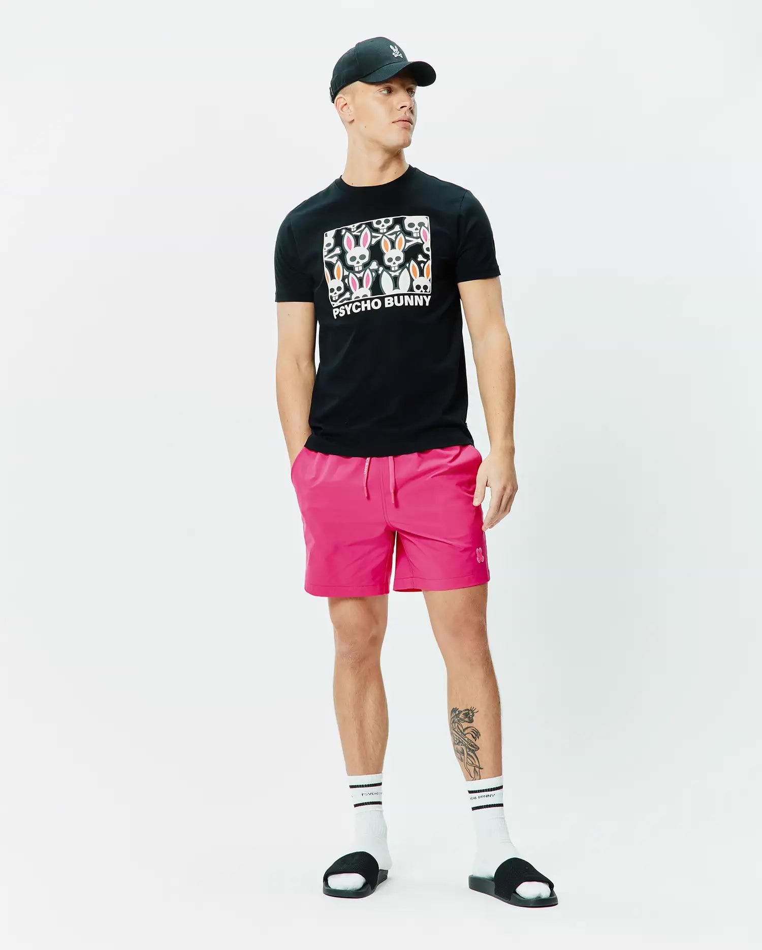 Mens Byron Graphic Tee, a black jersey t-shirt with a vibrant chest graphic of multiple Psycho Bunny skull logos and text, styled with bold hot pink drawstring shorts featuring a small embroidered bunny. (5)