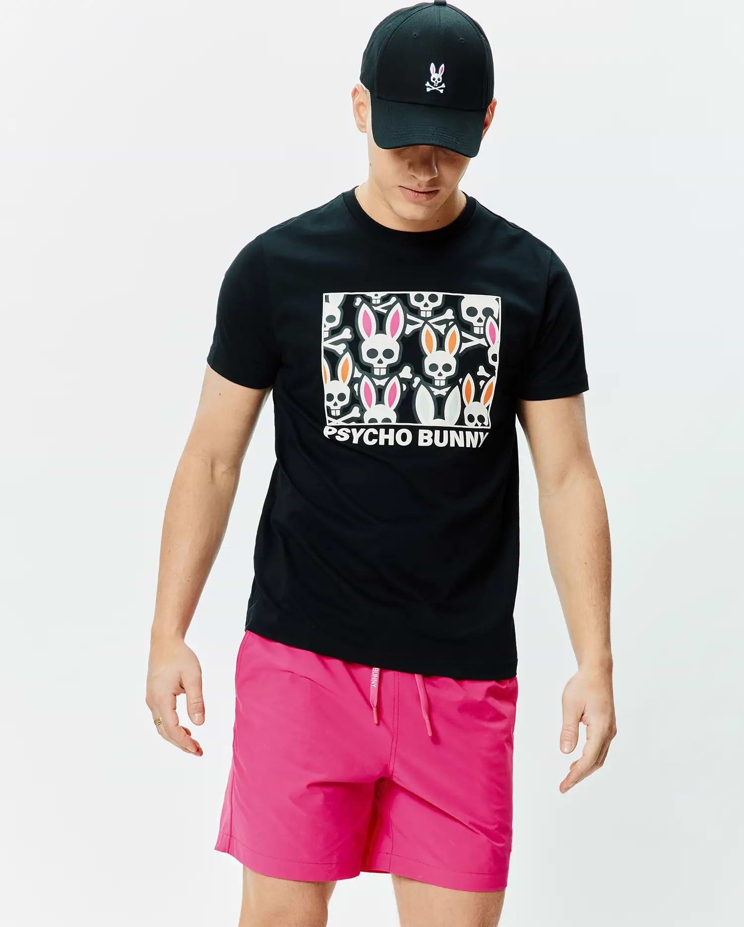 Black Mens Byron Graphic Tee with a large chest graphic of white bunny skulls, pink and orange ears, and Psycho Bunny text. Paired with bright pink drawstring shorts and a black cap with an embroidered Psycho Bunny logo. (4)