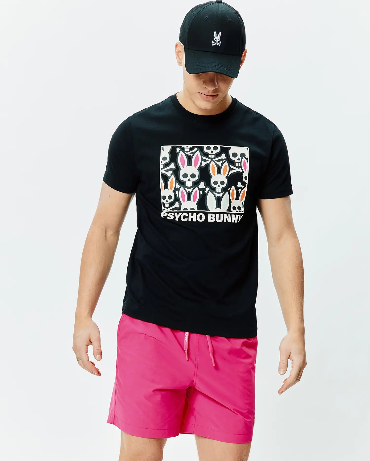 Black Mens Byron Graphic Tee with a large chest graphic of white bunny skulls, pink and orange ears, and Psycho Bunny text. Paired with bright pink drawstring shorts and a black cap with an embroidered Psycho Bunny logo. (4)
