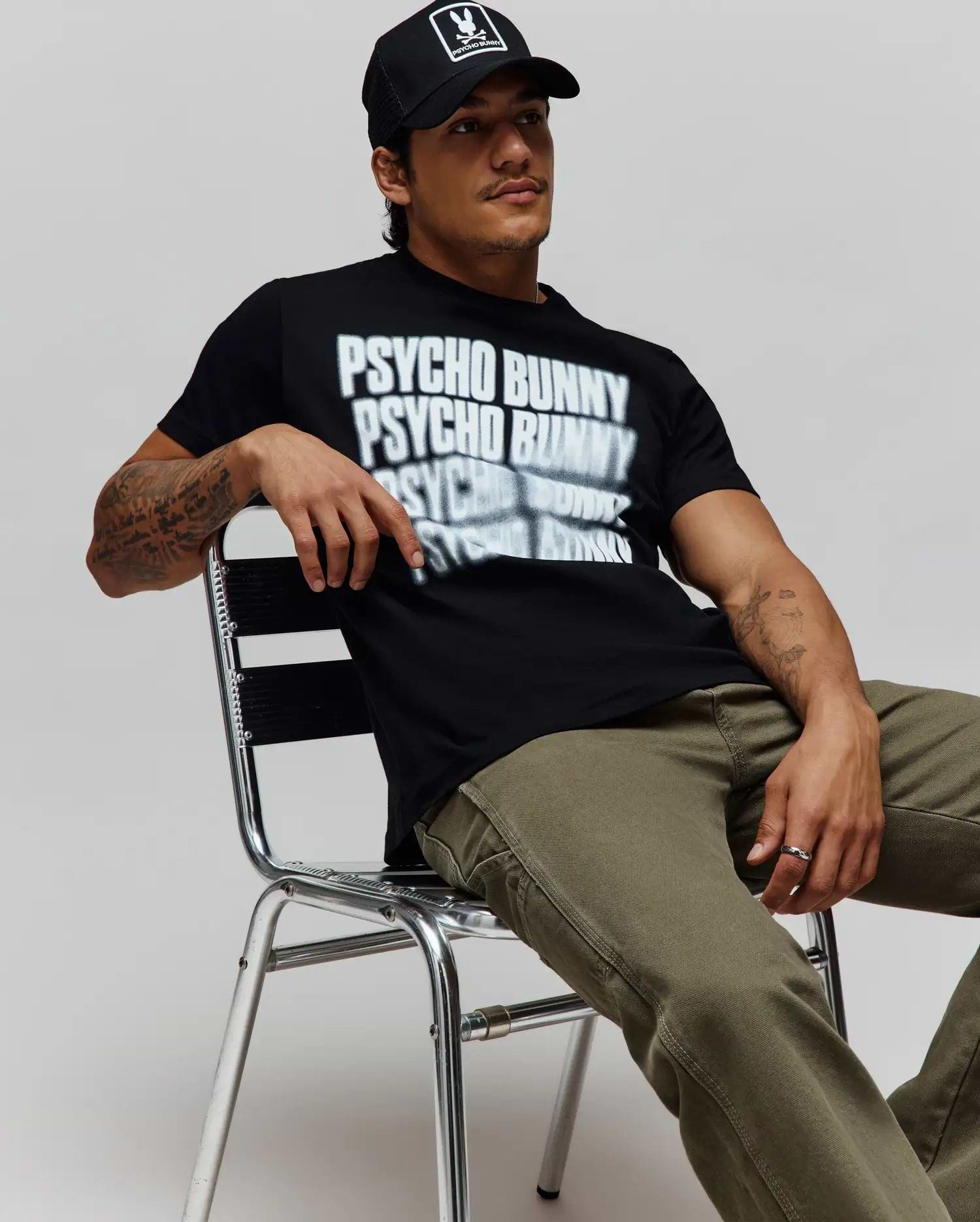 Black Mens Lawrence Graphic Tee, classic fit, with a large white blurred Psycho Bunny text graphic on soft jersey, paired with olive green canvas pants and a black cap featuring the iconic bunny skull logo. (4)