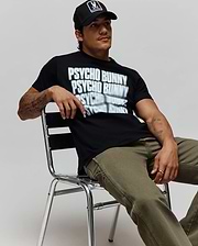 Black Mens Lawrence Graphic Tee, classic fit, with a large white blurred Psycho Bunny text graphic on soft jersey, paired with olive green canvas pants and a black cap featuring the iconic bunny skull logo. (4)