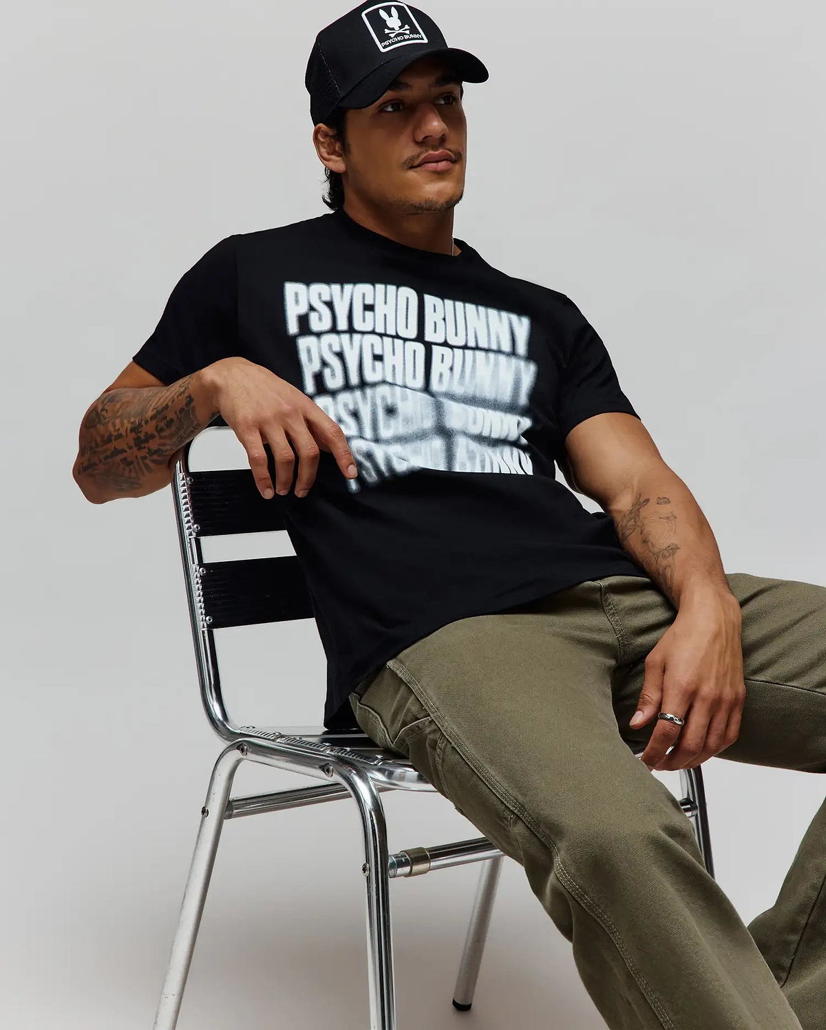 Black Mens Lawrence Graphic Tee, classic fit, with a large white blurred Psycho Bunny text graphic on soft jersey, paired with olive green canvas pants and a black cap featuring the iconic bunny skull logo. (4)