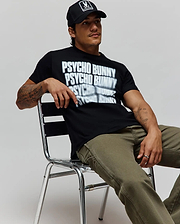Black Mens Lawrence Graphic Tee, classic fit, with a large white blurred Psycho Bunny text graphic on soft jersey, paired with olive green canvas pants and a black cap featuring the iconic bunny skull logo. (4)