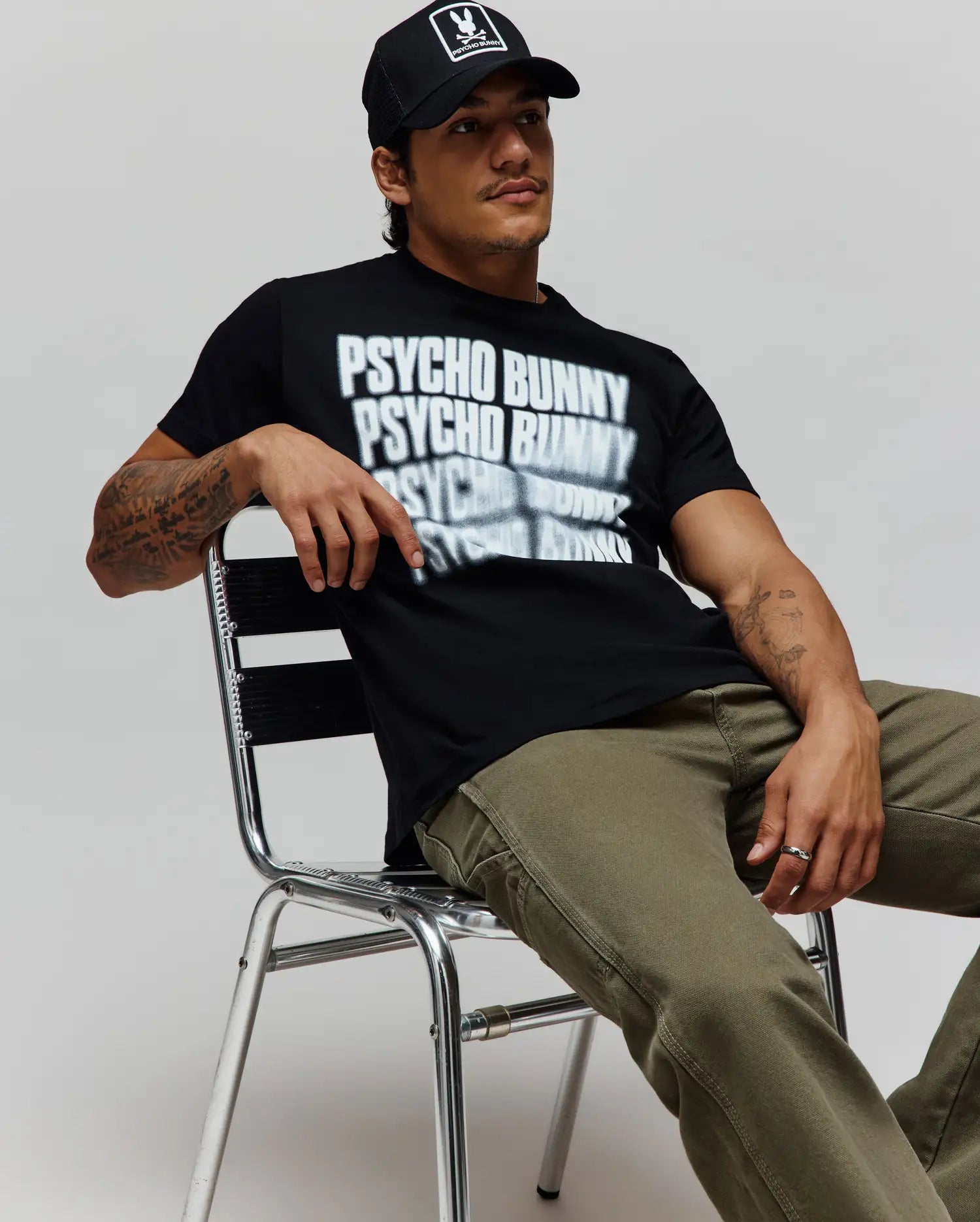Black Mens Lawrence Graphic Tee, classic fit, with a large white blurred Psycho Bunny text graphic on soft jersey, paired with olive green canvas pants and a black cap featuring the iconic bunny skull logo. (4)
