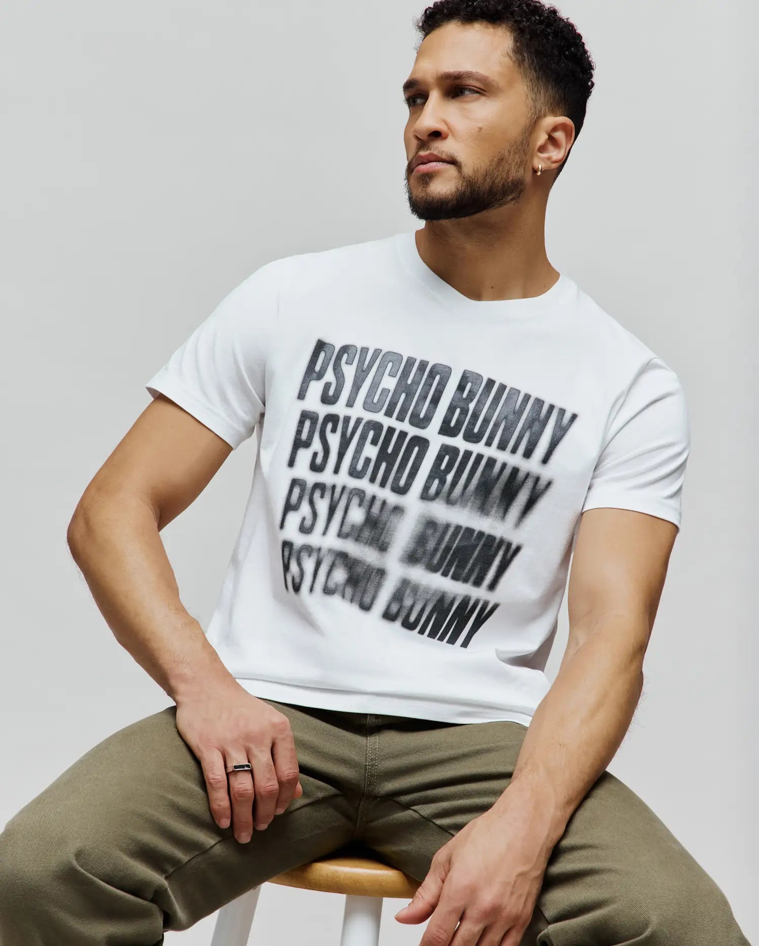 Mens Lawrence Graphic Tee, white classic fit jersey t-shirt featuring a large, central, blurred black and grey repeated Psycho Bunny text graphic. (4)