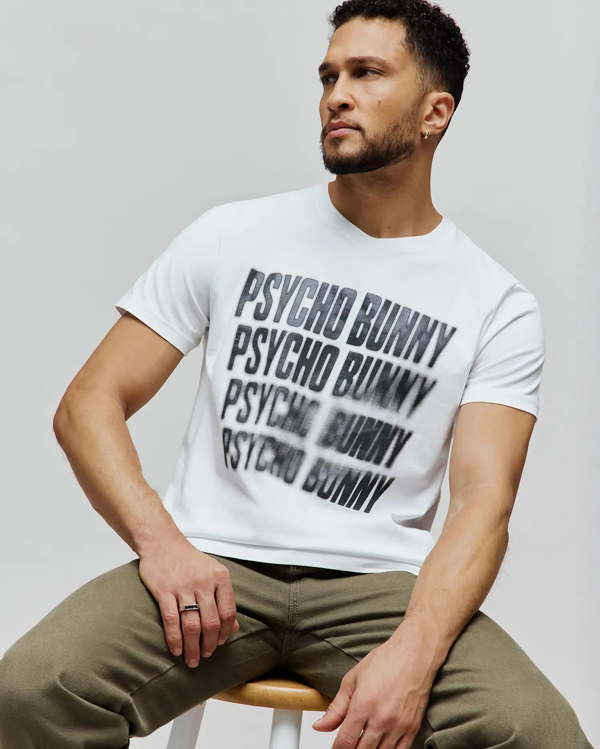 Mens Lawrence Graphic Tee, white classic fit jersey t-shirt featuring a large, central, blurred black and grey repeated Psycho Bunny text graphic. (4)