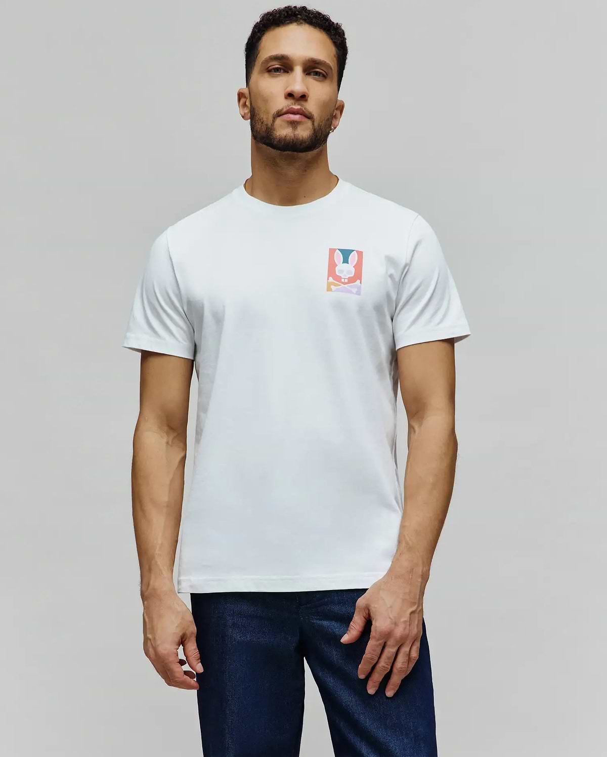 Mens Raphael Back Graphic Tee, a classic fit white jersey t-shirt featuring a small, colorful rectangular chest graphic of the iconic Psycho Bunny skull and crossbones logo in pink, blue, orange, and purple. (4)