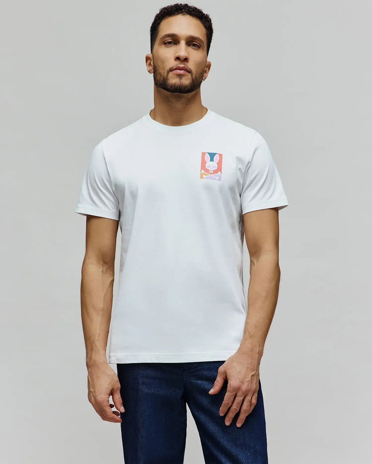 Mens Raphael Back Graphic Tee, a classic fit white jersey t-shirt featuring a small, colorful rectangular chest graphic of the iconic Psycho Bunny skull and crossbones logo in pink, blue, orange, and purple. (4)