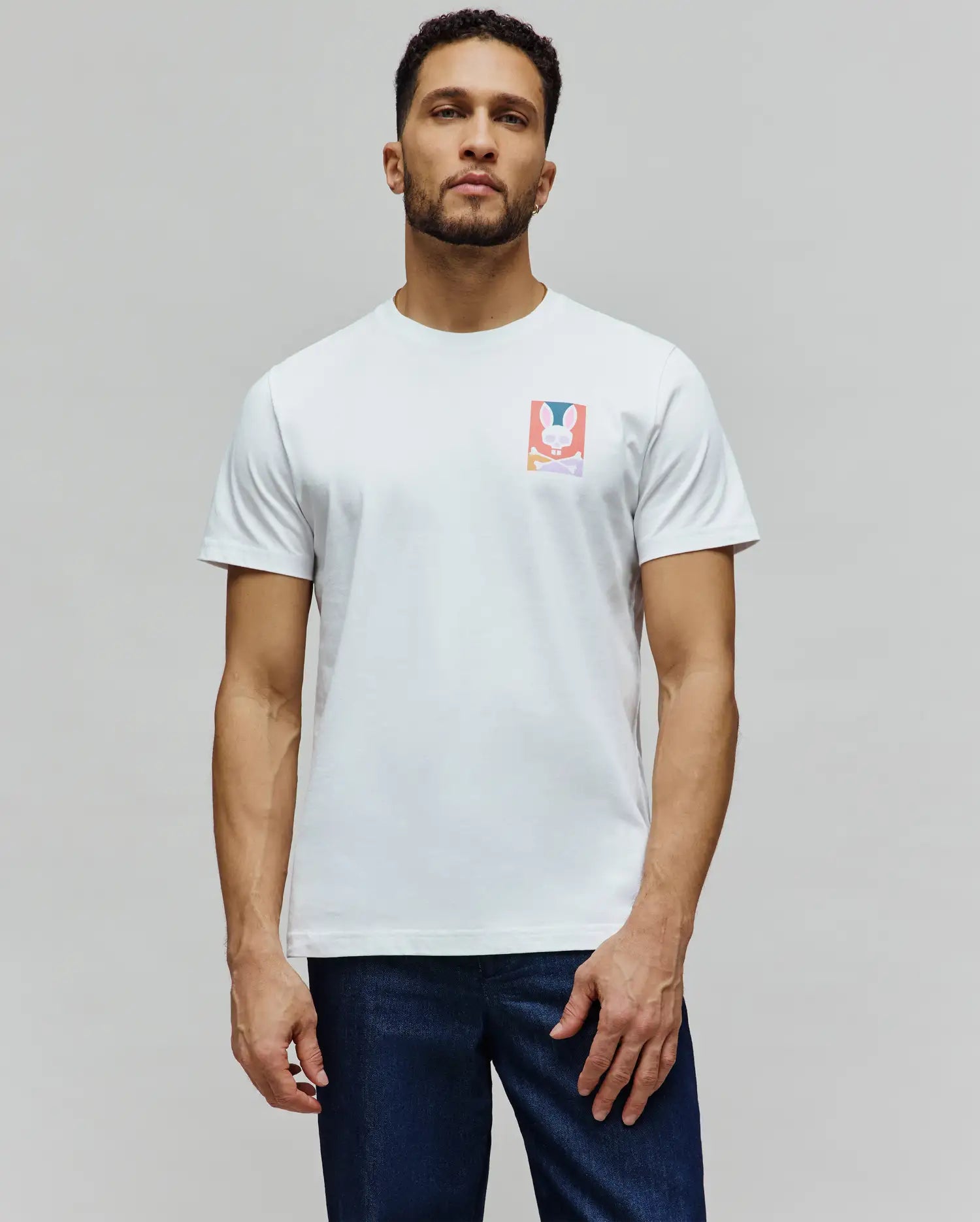 Mens Raphael Back Graphic Tee, a classic fit white jersey t-shirt featuring a small, colorful rectangular chest graphic of the iconic Psycho Bunny skull and crossbones logo in pink, blue, orange, and purple. (4)