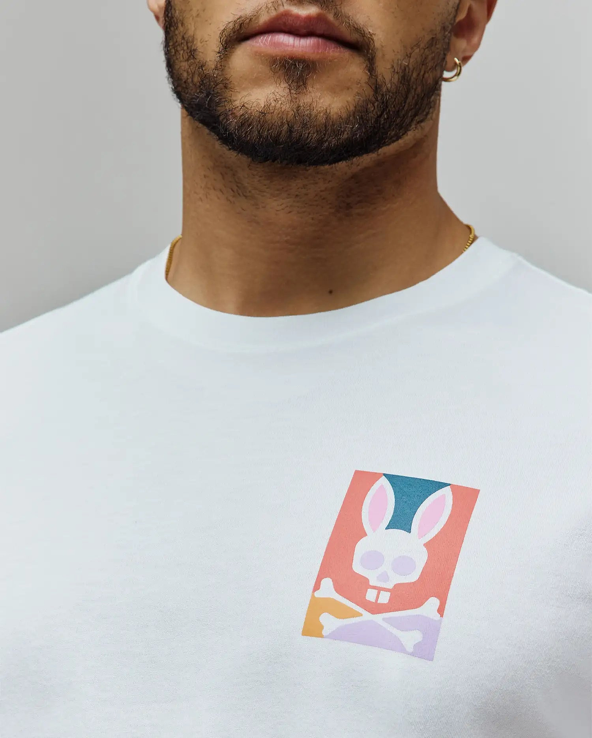 White Mens Raphael Back Graphic Tee, a classic fit jersey knit, featuring a small, vibrant chest graphic of the iconic Psycho Bunny skull logo in coral, teal, pink, and purple hues. (5)