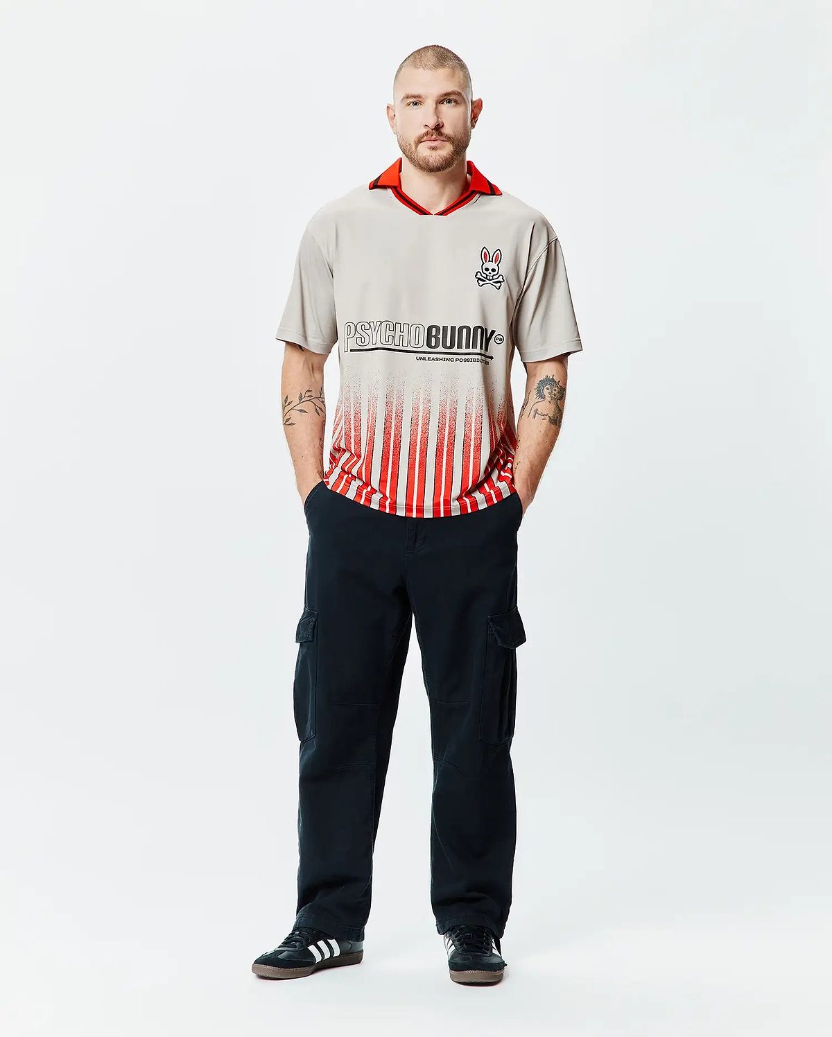 Mens Camden Oversized Soccer Jersey in stone grey with a vibrant red collar, featuring a large black Psycho Bunny text logo fading into bold red vertical stripes, and an embroidered bunny skull on the left chest. (4)