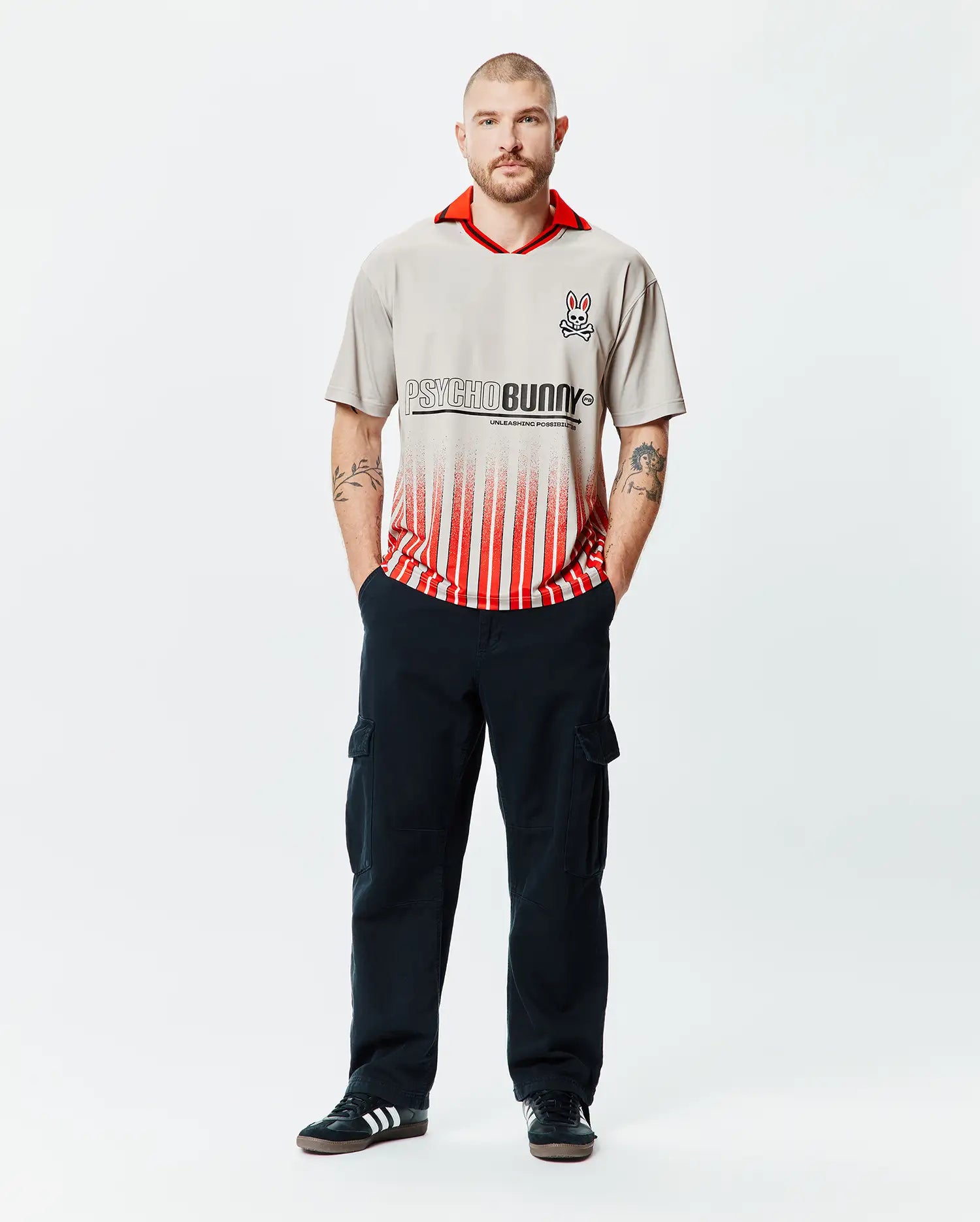 Mens Camden Oversized Soccer Jersey in stone grey with a vibrant red collar, featuring a large black Psycho Bunny text logo fading into bold red vertical stripes, and an embroidered bunny skull on the left chest. (4)