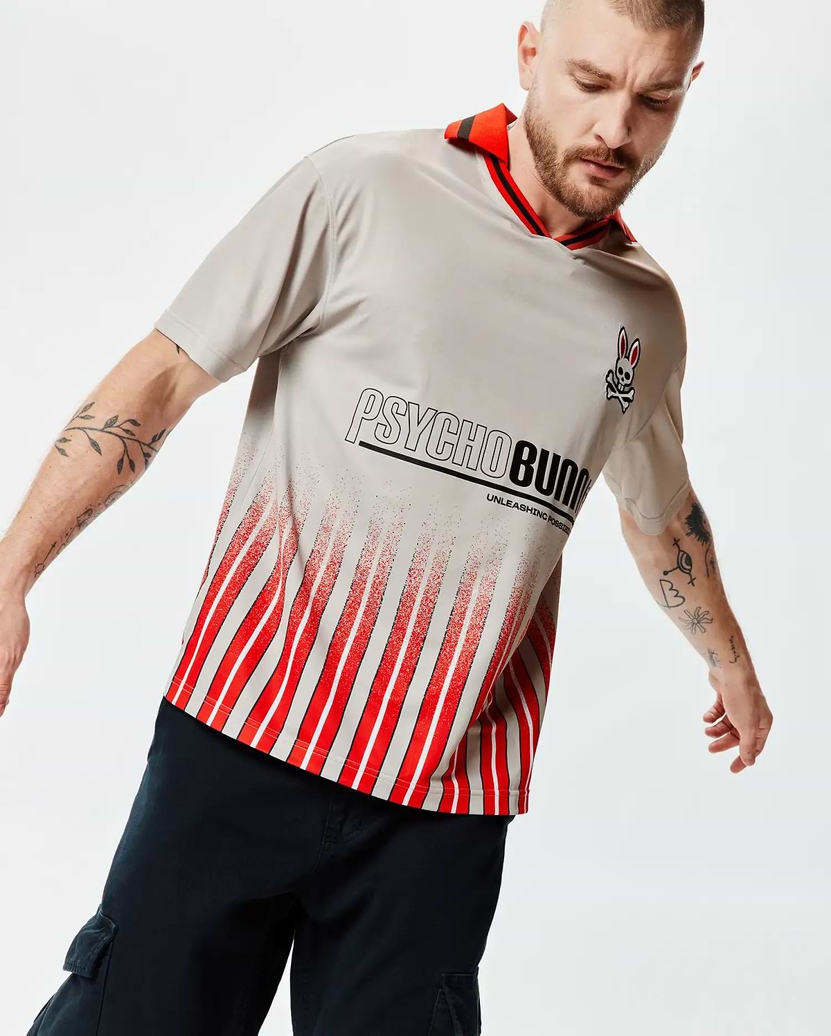 Mens Camden Oversized Soccer Jersey, light grey jersey knit, red striped collar, small Psycho Bunny skull logo, large Psycho Bunny text with Unleashing Possibilities, and a distressed red and white vertical stripe pattern. (3)