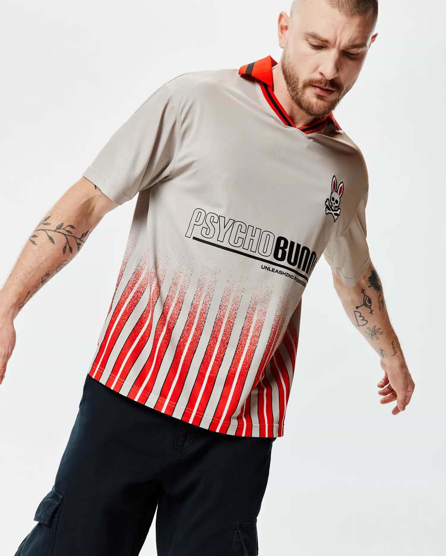 Mens Camden Oversized Soccer Jersey, light grey jersey knit, red striped collar, small Psycho Bunny skull logo, large Psycho Bunny text with Unleashing Possibilities, and a distressed red and white vertical stripe pattern. (3)